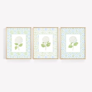 May include: A set of three framed wall art prints, each featuring a white hydrangea flower centred on a white background, surrounded by a light blue and green damask pattern border, all enclosed in thin gold-toned rectangular frames.