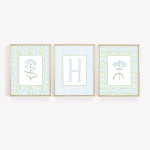 May include: Three rectangular wall art prints in thin gold frames. The outer two prints feature light blue floral illustrations on a pale green and blue damask background. The center print displays a large light blue letter H on a blue striped background.