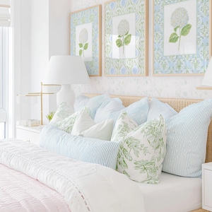 May include: A bedroom scene featuring a woven rattan headboard, a light pink quilted bedspread, and an assortment of pillows in blue striped and green botanical patterns. Three framed floral prints hang above the bed on a white wall.