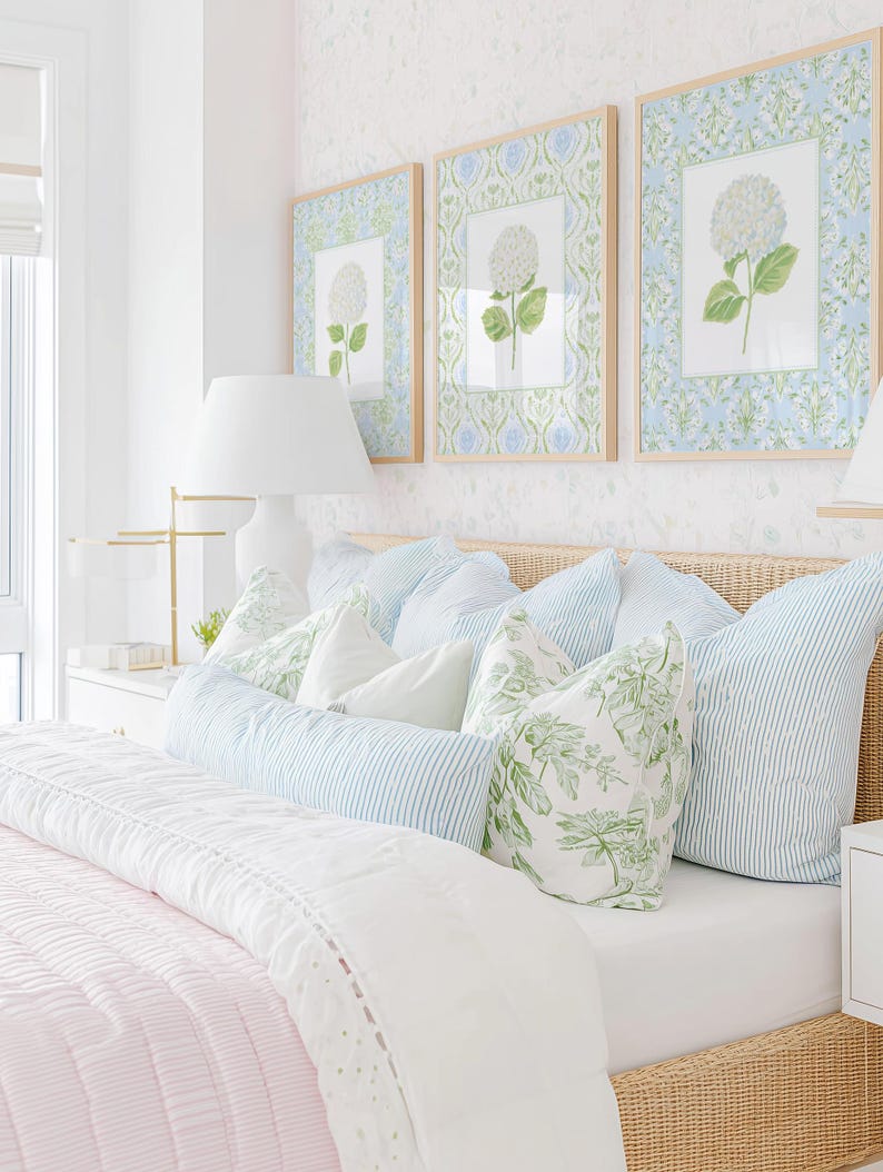 May include: A bedroom scene featuring a woven rattan headboard, a light pink quilted bedspread, and an assortment of pillows in blue striped and green botanical patterns. Three framed floral prints hang above the bed on a white wall.