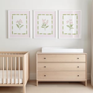 May include: Three framed floral prints with pink gingham backgrounds and green borders hang above a light wood dresser with black knobs. A light wood crib is to the left.