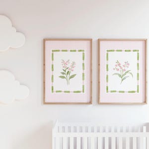 May include: Two framed floral art prints. Each print features a watercolor painting of pink flowers with green leaves, set against a pink gingham background. The frames are a light brown color. White cloud-shaped wall decor is also visible.