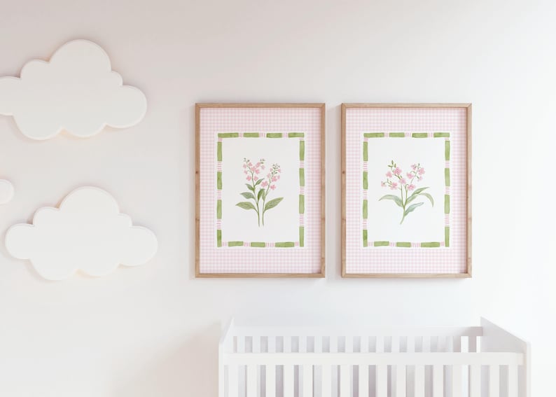 May include: Two framed floral art prints. Each print features a watercolor painting of pink flowers with green leaves, set against a pink gingham background. The frames are a light brown color. White cloud-shaped wall decor is also visible.