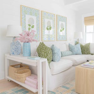 May include: A white console table with two woven baskets and two pink books on the lower shelf. On top sits a blue patterned lamp, a blue striped vase with pink flowers, and a light blue book. Three framed floral prints hang on the wall.