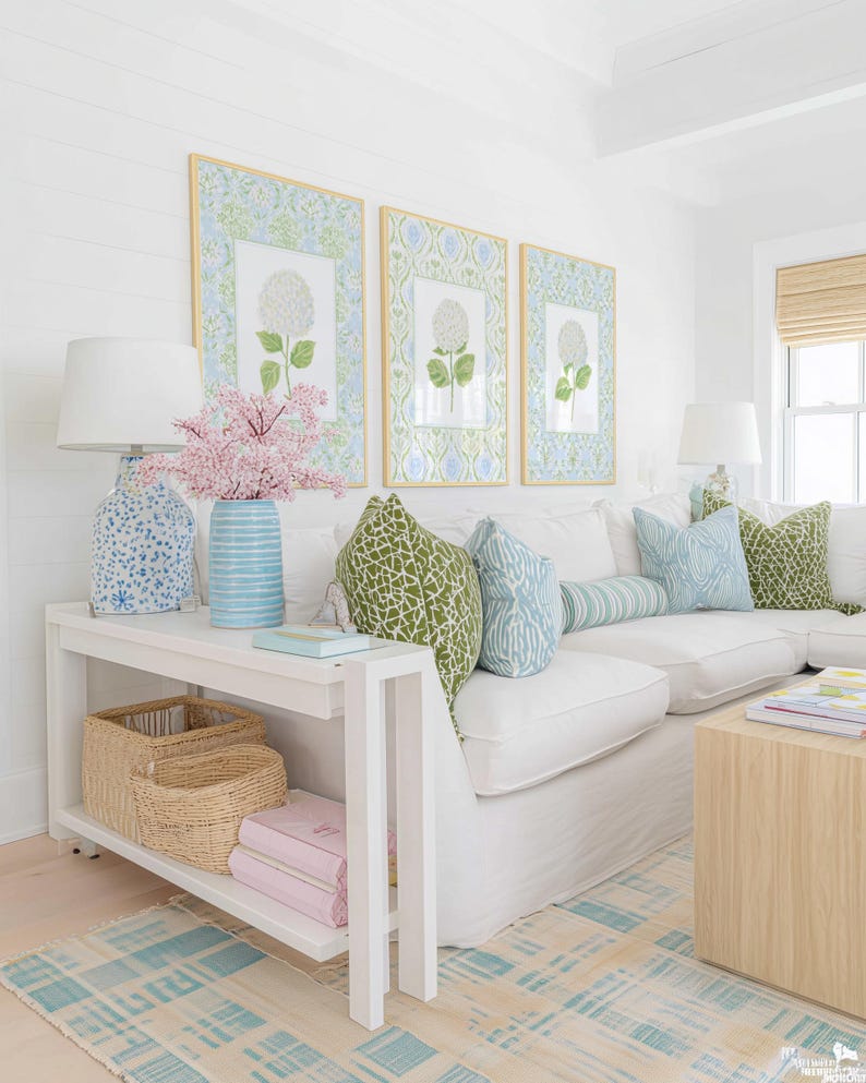 May include: A white console table with two woven baskets and two pink books on the lower shelf. On top sits a blue patterned lamp, a blue striped vase with pink flowers, and a light blue book. Three framed floral prints hang on the wall.