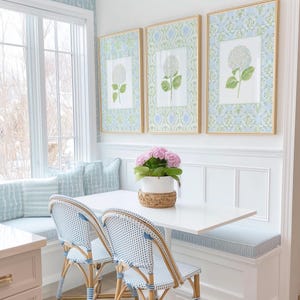 May include: A dining nook featuring a white rectangular table, two bistro chairs with blue and white woven patterns, and a built-in bench with light blue striped cushions. Three framed botanical prints of white hydrangeas hang on the wall.