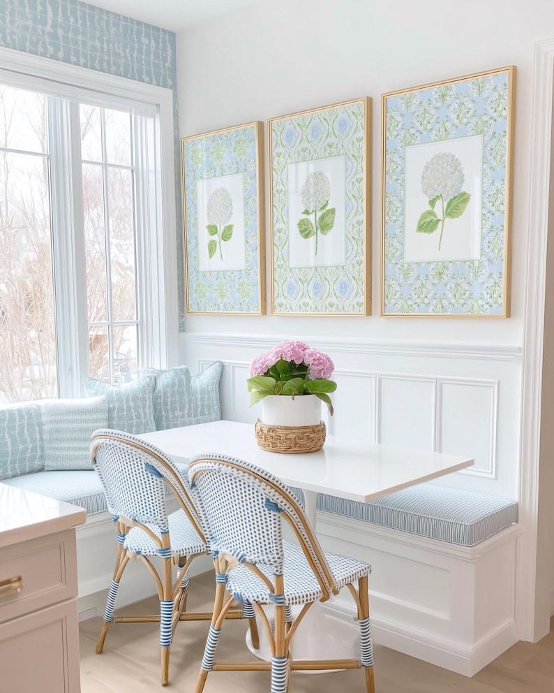 May include: A dining nook featuring a white rectangular table, two bistro chairs with blue and white woven patterns, and a built-in bench with light blue striped cushions. Three framed botanical prints of white hydrangeas hang on the wall.