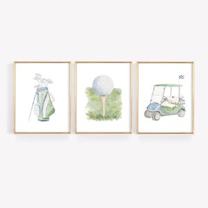 May include: A set of three framed watercolour prints with golf-themed illustrations. The prints show a golf bag with clubs, a golf ball on a tee, and a golf cart. The colour scheme includes shades of green, blue, and white.