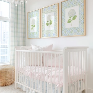 May include: A white cot with a light pink mattress cover and a blue and white plaid ruffled skirt. Above the cot are three framed prints featuring white hydrangea illustrations on a blue and green patterned background in gold frames.