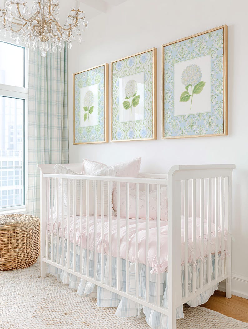 May include: A white cot with a light pink mattress cover and a blue and white plaid ruffled skirt. Above the cot are three framed prints featuring white hydrangea illustrations on a blue and green patterned background in gold frames.