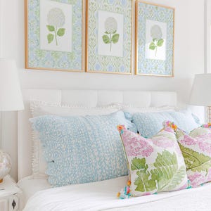 May include: Three framed botanical prints featuring white hydrangea illustrations on patterned backgrounds, displayed above a white upholstered headboard with light blue patterned pillows and decorative cushions with green leaf prints.