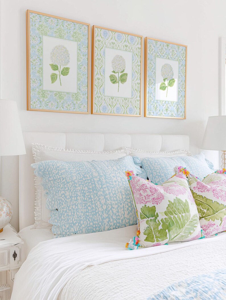 May include: Three framed botanical prints featuring white hydrangea illustrations on patterned backgrounds, displayed above a white upholstered headboard with light blue patterned pillows and decorative cushions with green leaf prints.