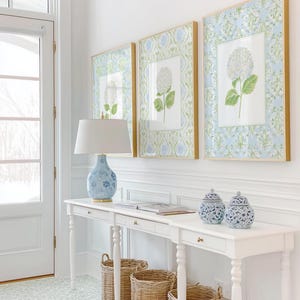 May include: A white console table with three drawers, topped with a blue and white patterned lamp and two matching lidded jars. Three framed botanical prints of white hydrangeas hang above, while three woven wicker baskets sit underneath.