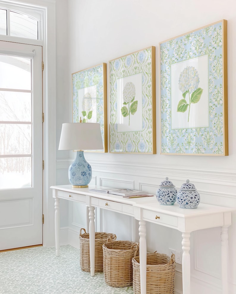 May include: A white console table with three drawers, topped with a blue and white patterned lamp and two matching lidded jars. Three framed botanical prints of white hydrangeas hang above, while three woven wicker baskets sit underneath.