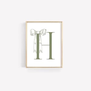 May include: A framed print featuring the letter "H" in a muted green hue, adorned with a detailed bow illustration. The bow and letter are set against a white background, encased in a gold frame, creating a classic and elegant aesthetic.