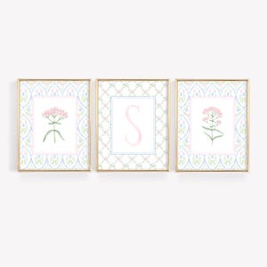 May include: Three framed floral art prints. Each print has a gold frame and features a floral design in pink and green, set against a light blue and white patterned background. The center print displays the letter "S".