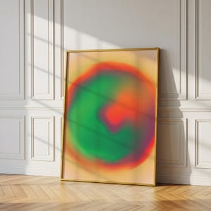 May include: A framed abstract art print featuring a blurred, colorful design. The artwork showcases a central green and red shape against a light yellow background. The frame is gold, and the print leans against a white wall on a wooden floor.