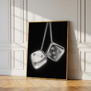 May include: A framed print showing two silver dice hanging from chains against a black backdrop. The dice have visible dots and reflect light. The frame is gold, and the artwork leans against a white wall and wooden floor.