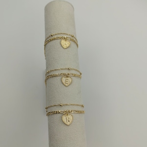 May include: Three gold chain bracelets with heart-shaped charms. Each charm has a different letter, H, S, and R, in the center.