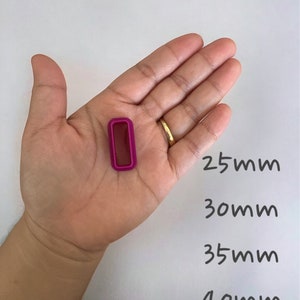 May include: A close-up of a hand holding a small, rectangular, fuchsia-colored object. The object is about 2.5 cm wide and 4 cm long. The hand is holding the object in front of a white background with the measurements 25mm, 30mm, 35mm, and 40mm written below the object.