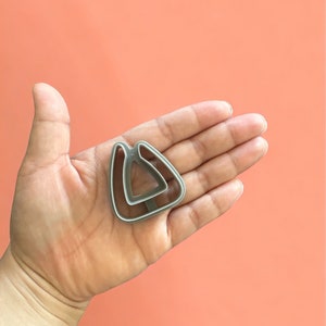 May include: A silver-colored metal cookie cutter in the shape of a triangle with rounded corners and a handle. The cutter is held in an open hand against a peach-colored background.