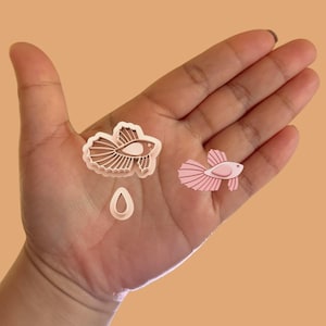 May include: Two cookie cutters in the shape of a betta fish. One is a light brown color and the other is pink. A small, light brown cookie cutter in the shape of a water drop is also included.