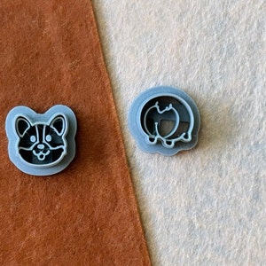 May include: Two gray plastic cookie cutters, one shaped like a corgi dog and the other shaped like a pumpkin.