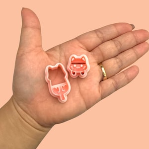 May include: Two pink plastic cookie cutters, one shaped like a cat and the other shaped like a cat holding a popsicle.