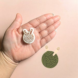 May include: White plastic cookie cutter shaped like a ball of yarn with two knitting needles sticking out. The cookie cutter is being held in a hand.