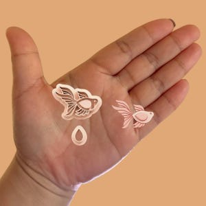 May include: Two pink plastic cookie cutters shaped like fish, one larger than the other, and a smaller pink plastic cookie cutter shaped like a teardrop. The cookie cutters are on a person's hand.