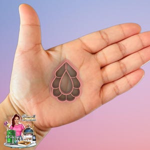 May include: A pink teardrop-shaped cookie cutter with a layered design. The cutter is held in a hand against a gradient background. The cutter's design features a central teardrop shape surrounded by layered, petal-like forms.