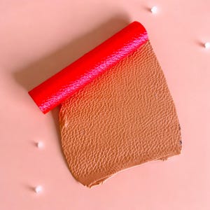 May include: A piece of brown leather with a textured surface is rolled up with a bright red tube. The leather is on a light pink background.