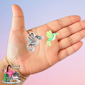 May include: Two hummingbird-themed items. One is a gray plastic cookie cutter, and the other is a green and white hummingbird-shaped embellishment. The items are displayed on a person's open palm.