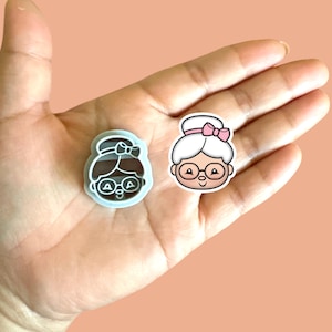 May include: A white and pink cookie cutter in the shape of a cartoon grandma with glasses and a bun. The cookie cutter is on a hand.