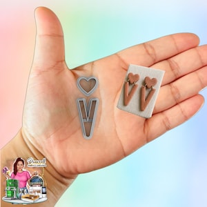 May include: A hand displays a silver metal heart and V-shaped cookie cutter, alongside a pair of brown clay earrings with heart and V shapes. The earrings are on a small, rectangular, beige card.
