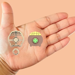 May include: A set of three cookie cutters in the shape of a pot of gold, a four-leaf clover, and a single leaf clover. The pot of gold cookie cutter is brown with yellow coins and a green four-leaf clover on top. The cookie cutters are made of plastic.