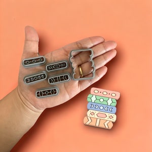 May include: A set of seven gray plastic cookie cutters with geometric designs. The cutters are arranged on a hand, and a stack of five colorful cookies with the same geometric designs is shown to the right.