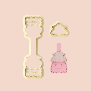 May include: Three cookie cutters in a light yellow color. One is a tall, ornate shape, another is a simple house outline, and the third is a pink and tan mushroom shape with a smiling face. All are on a light pink background.