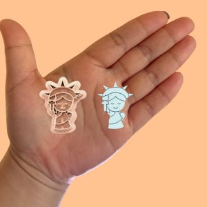 May include: Two cookie cutters in the shape of a cartoon person dressed as the Statue of Liberty. One cutter is brown and the other is light blue.