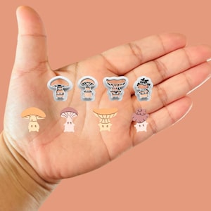 May include: A hand holding a collection of mushroom-shaped cookie cutters and mushroom-shaped cookies. The cutters are silver and the cookies are in shades of orange, pink, and brown. The background is a solid peach color.