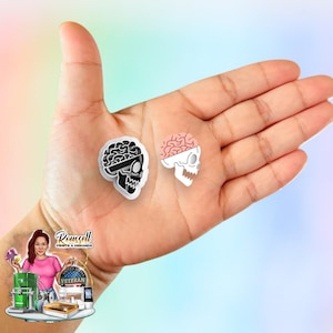 May include: Two skull-shaped stickers. One is black and white with a brain design, the other is white with a pink brain design. The stickers are held in a hand against a pastel background. The stickers are likely for crafting or scrapbooking.