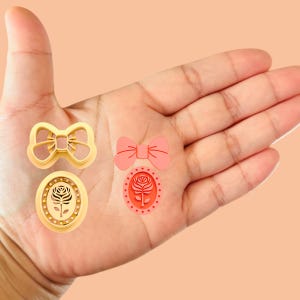 May include: Four cookie cutters in a hand. Two are gold and shaped like bows. One is pink and shaped like a bow. The last is pink and oval with a rose design.