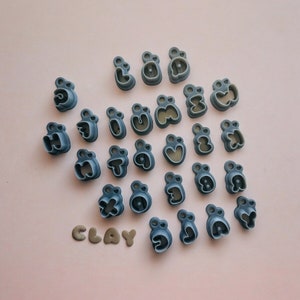 May include: A set of 26 gray plastic alphabet cookie cutters. The cutters are arranged in a semi-circle on a light pink background. The word "CLAY" is written in gray letters below the cutters.