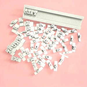 May include: A set of white plastic letter and number stamps, scattered on a pink surface. The stamps are arranged in a random pattern, with some of the letters and numbers visible. The stamps are used for creating custom labels and tags.