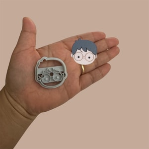 May include: A gray plastic cookie cutter in the shape of a cartoon boy with black hair and glasses. The cookie cutter is being held in a person's hand.