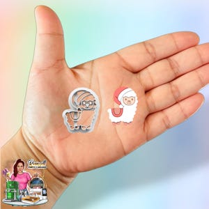 May include: Two llama-shaped cookie cutters. One is a tan llama wearing glasses and a hat. The other is a white llama wearing a red Santa hat. The cutters are held in a hand.