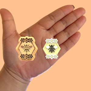 May include: Two gold-colored bee-themed pins. One pin features a bee inside a honeycomb with a floral border. The other pin features a bee inside a honeycomb with a daisy border.