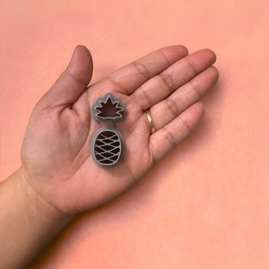 May include: Two gray pineapple-shaped cookie cutters. One cutter is the pineapple crown, and the other is the pineapple body. The cutters are made of plastic and are on a pink background.