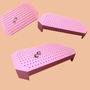 May include: Three pink earring display boards with multiple holes. Two pairs of earrings are visible on two of the boards. The boards have a rectangular shape with a slightly angled top and are elevated by short legs.