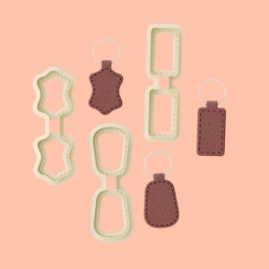 May include: Five leather keychains with different shapes and sizes. The keychains are brown and have a stitched edge. The keychains are hanging from metal rings.